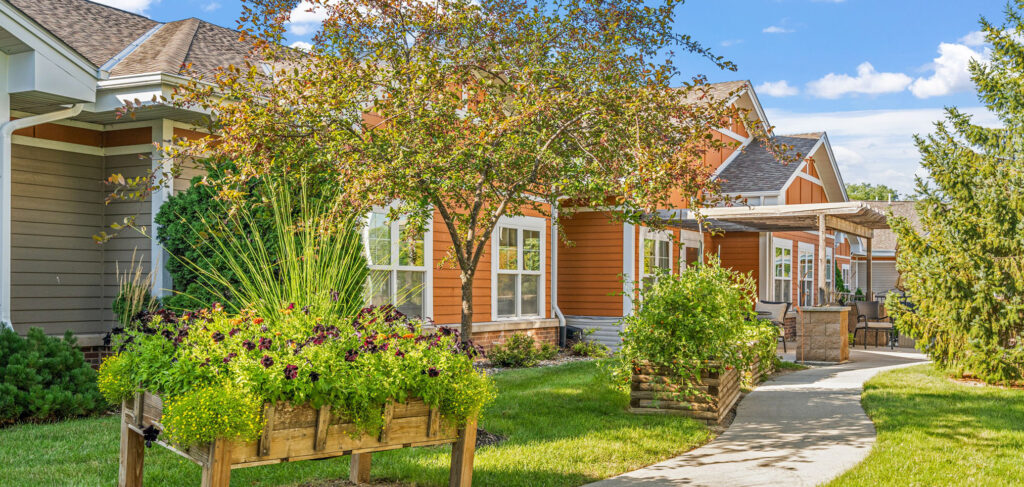 This is a picture of Newton Village's garden, featuring raised planters with vibrant flowers, a winding concrete path and a seating area under a pergola. The building has warm-toned siding and large windows.